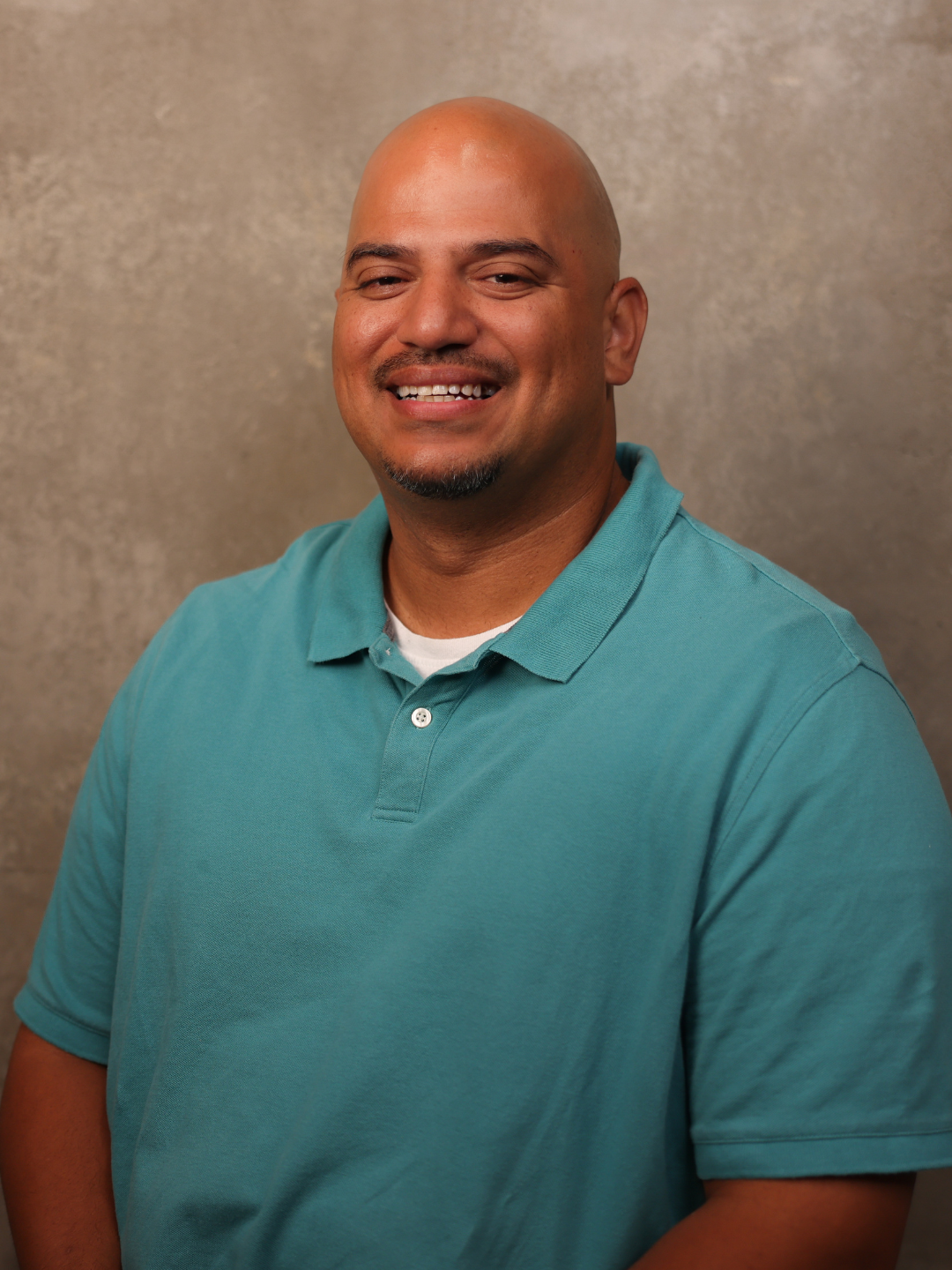 Portrait of a smiling bald man with a goatee wearing a teal polo shirt against a textured beige background.
