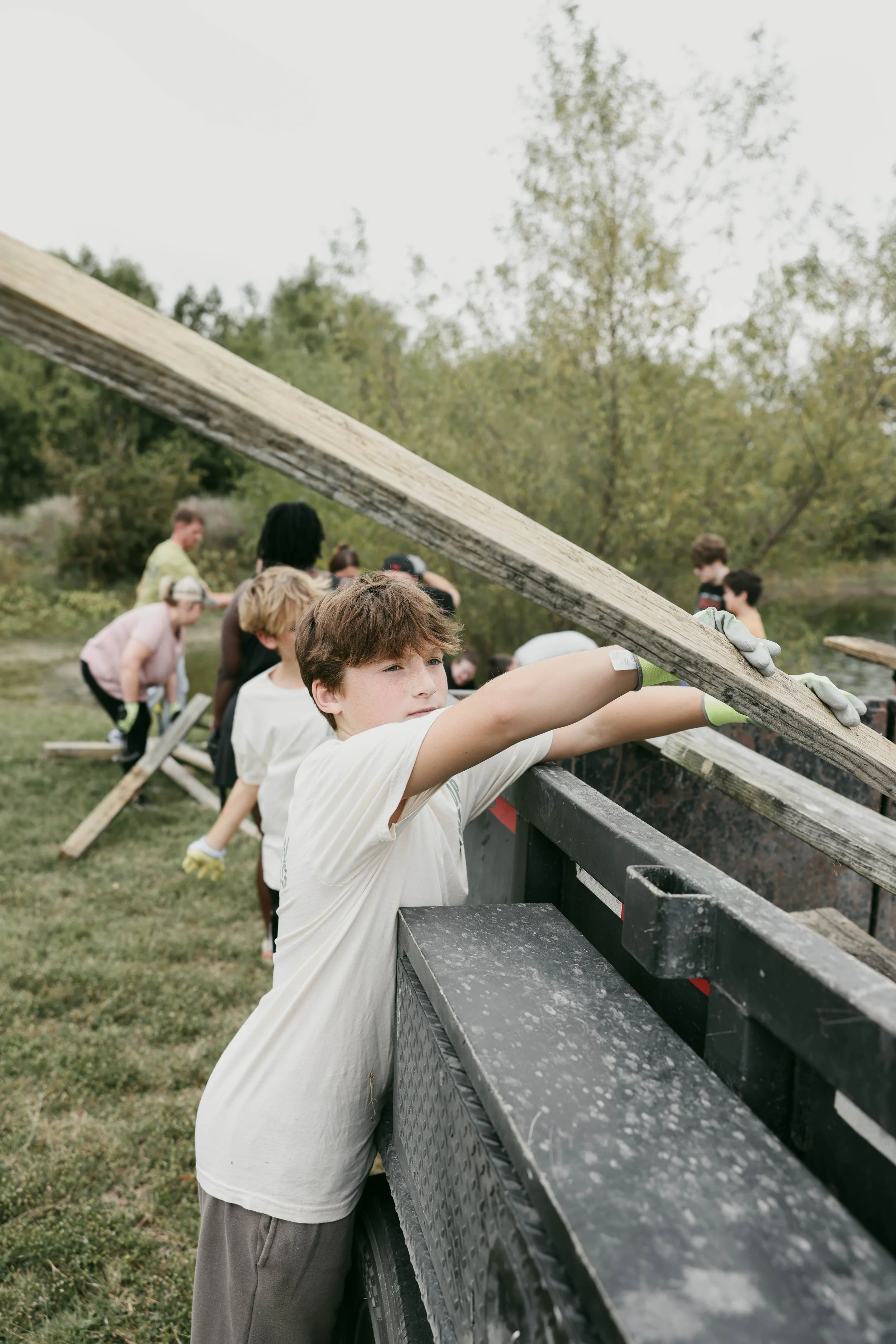 A group of children standing on the back of a flatbed truck, holding and positioning large wooden planks during an outdoor activity or project in a grassy area with trees in the background.
