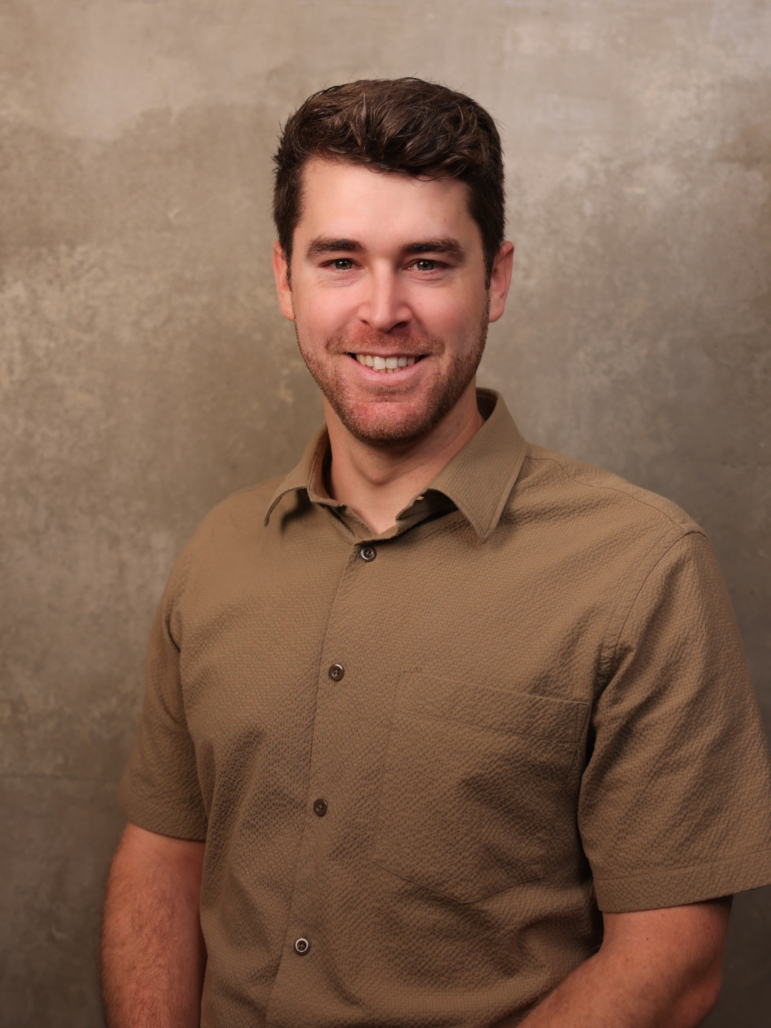 A smiling young man with dark wavy hair and facial hair, wearing a brown button-up shirt, standing against a textured beige background.