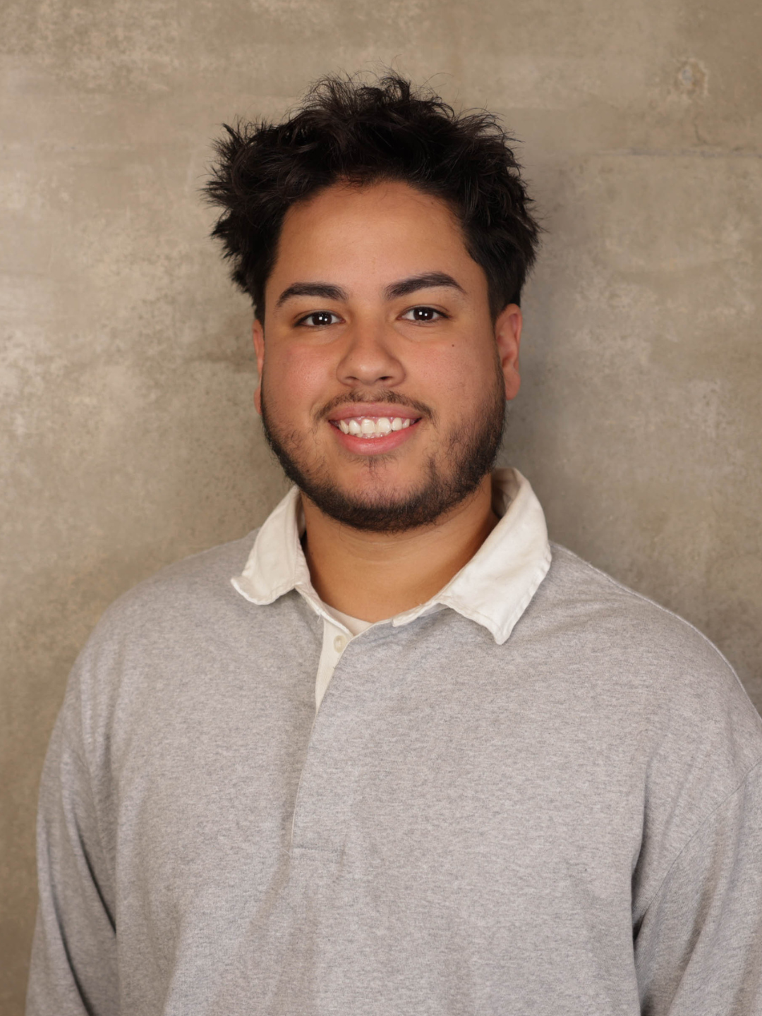 Portrait of a young man with dark, curly hair and a beard smiling, wearing a light grey collared sweater over a white shirt, standing against a beige textured wall.