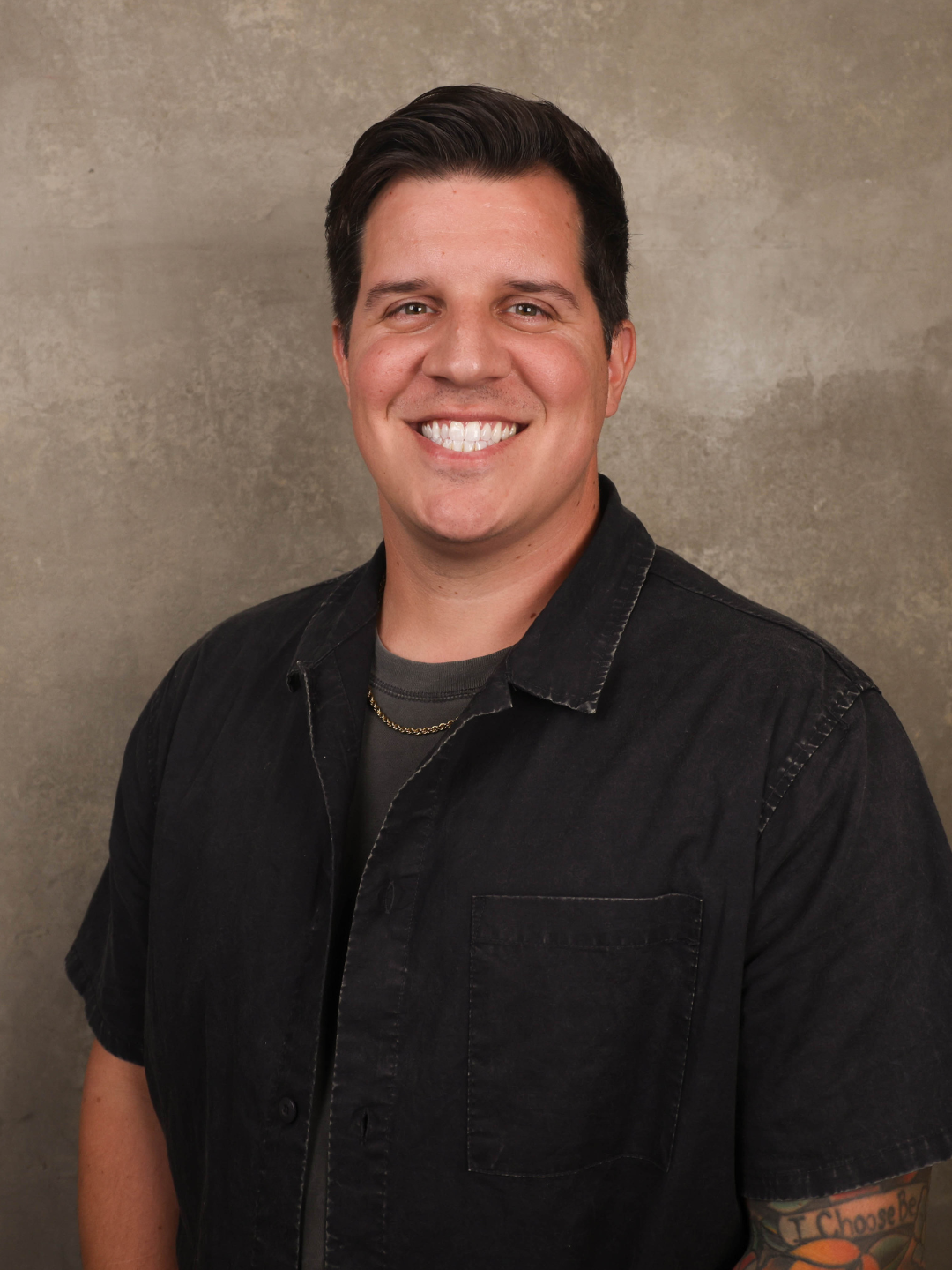 Portrait of a smiling man with dark hair, wearing a black shirt and a gold chain, standing against a beige background.