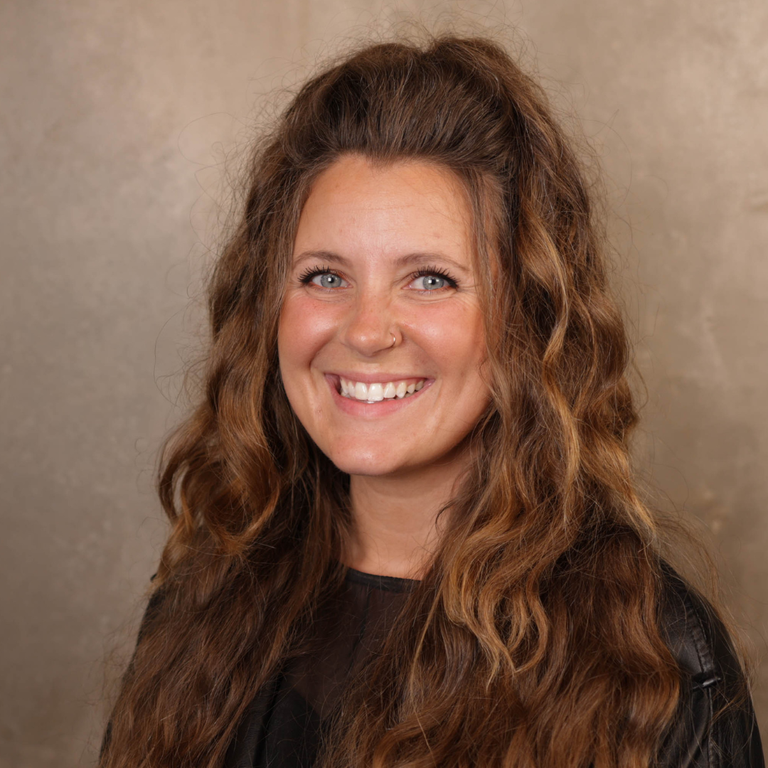 Portrait of a woman with long wavy brown hair, blue eyes, and a nose piercing, smiling against a neutral background.