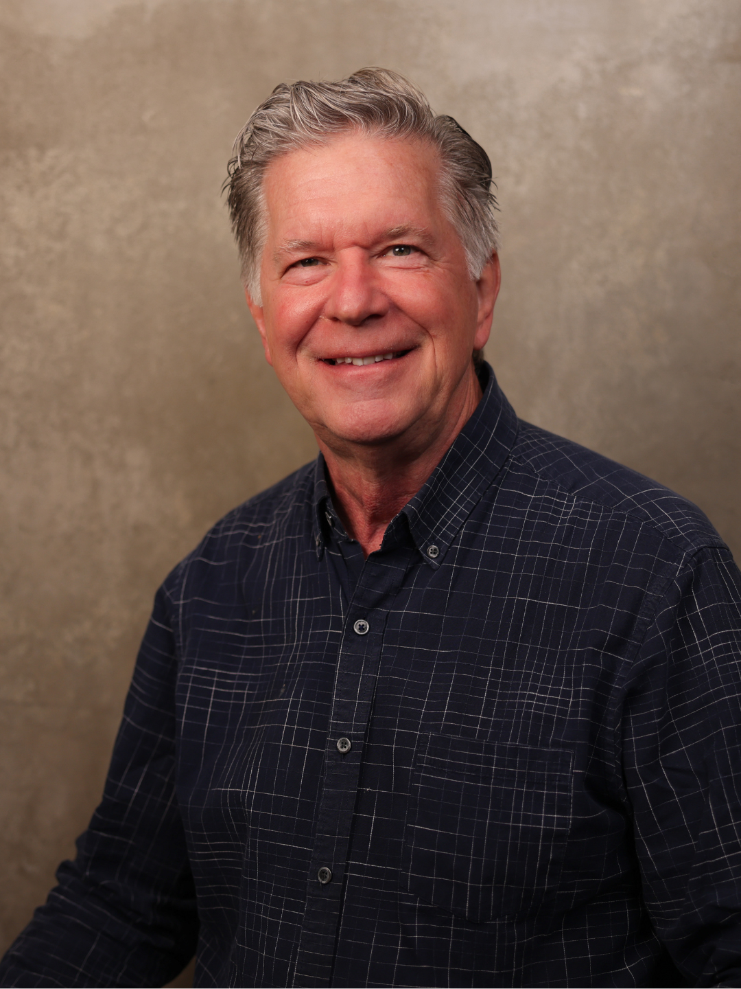 Portrait of a smiling older man with gray hair, wearing a dark checkered shirt, standing against a beige textured background.
