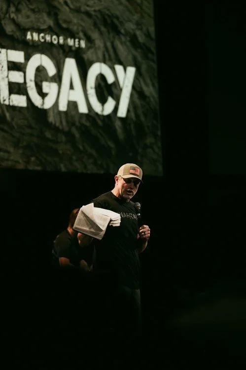 A man wearing a baseball cap and black shirt holds a towel and a microphone on a dark stage, with a large screen behind him displaying the words 'ANCHOR MEN LEGACY'.