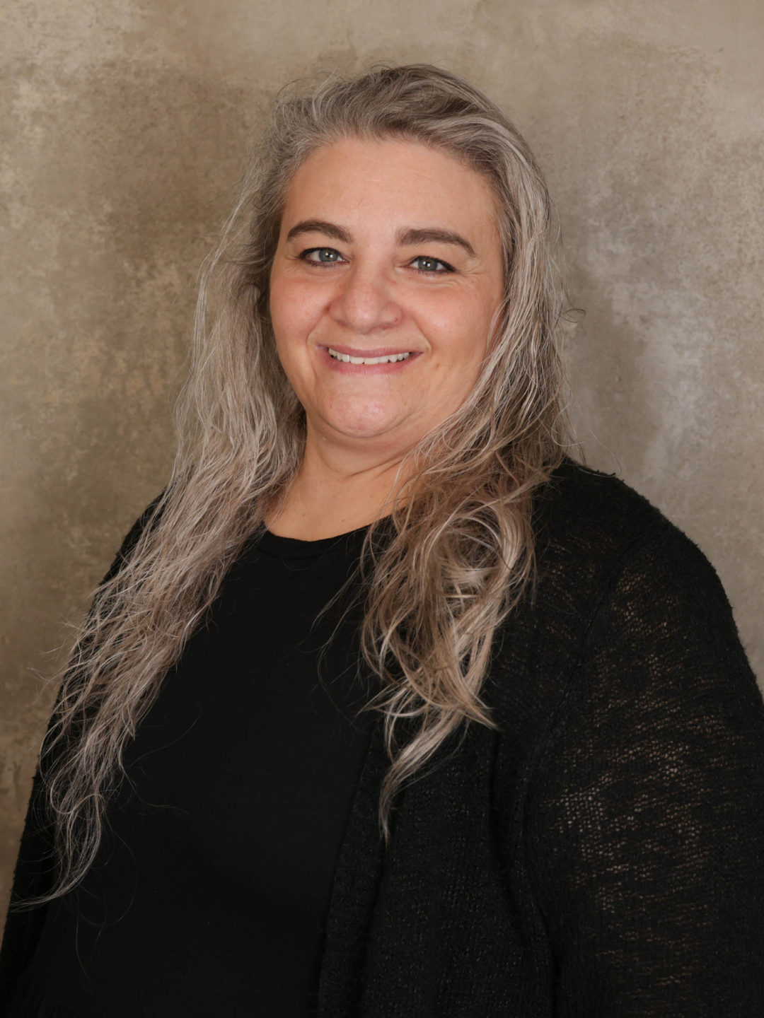 A woman with long, curly, gray-blonde hair smiling, wearing a black top and black cardigan, standing against a beige background.