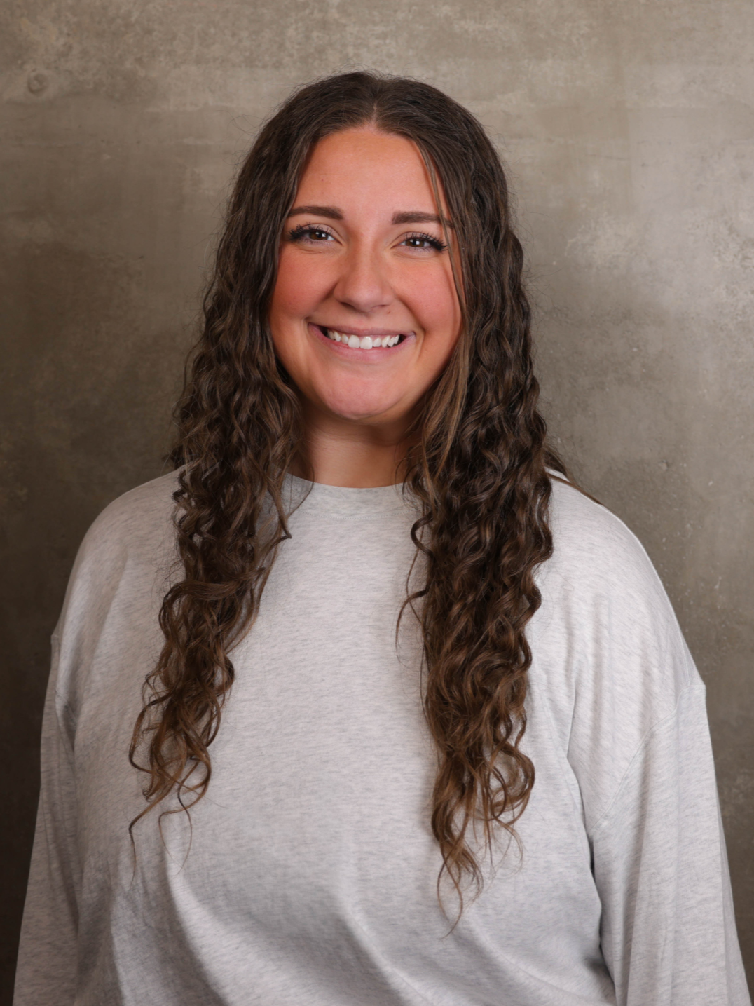 A young woman with long, curly brown hair smiling in front of a gray textured wall, wearing a light gray sweatshirt.