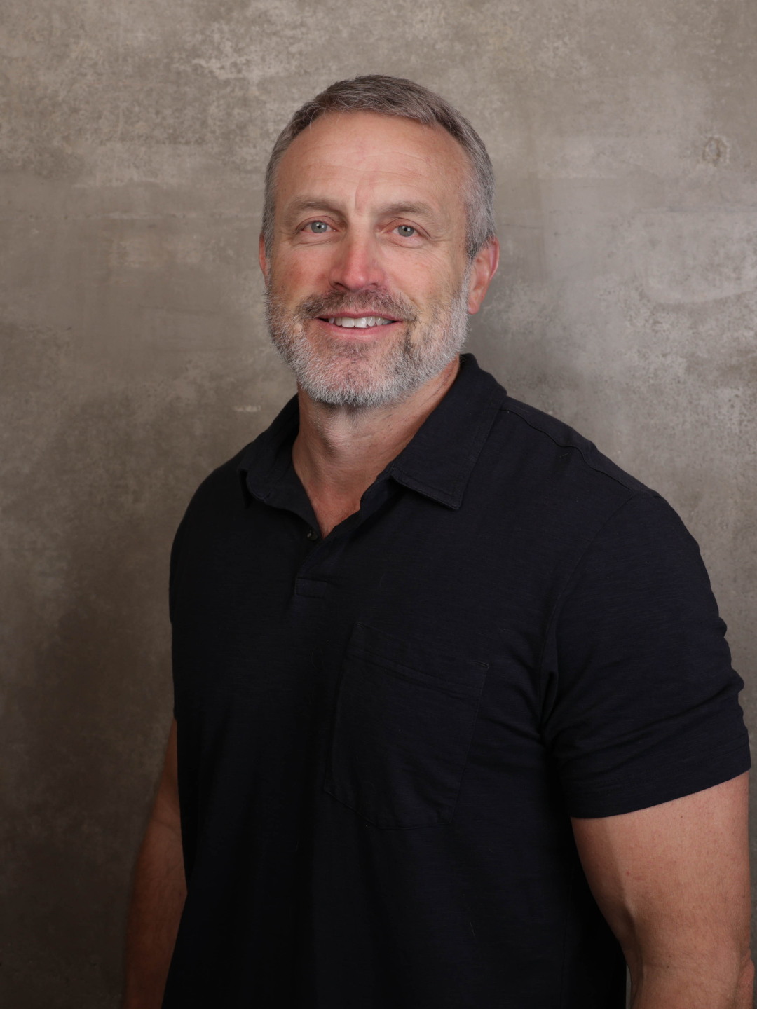 A middle-aged man with gray hair and a beard, wearing a black polo shirt, smiling against a textured concrete wall background.