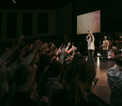 A church service with a preacher on stage addressing a congregation with raised hands, in a darkened room with a large screen behind.