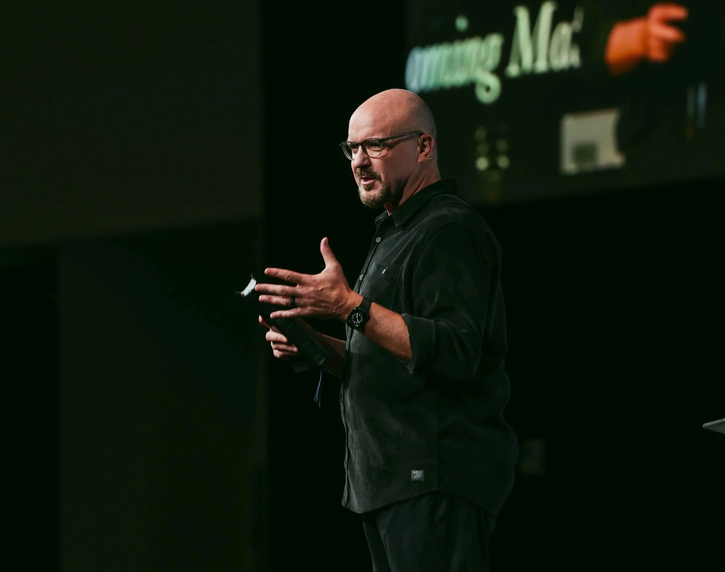 A man with glasses and a bald head speaking or presenting on stage, dressed in black, with a dark background and a screen displaying the words 'Church' and a pointing arrow.