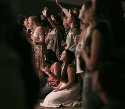 People at a concert or religious gathering, some standing with hands raised, others kneeling and praying.