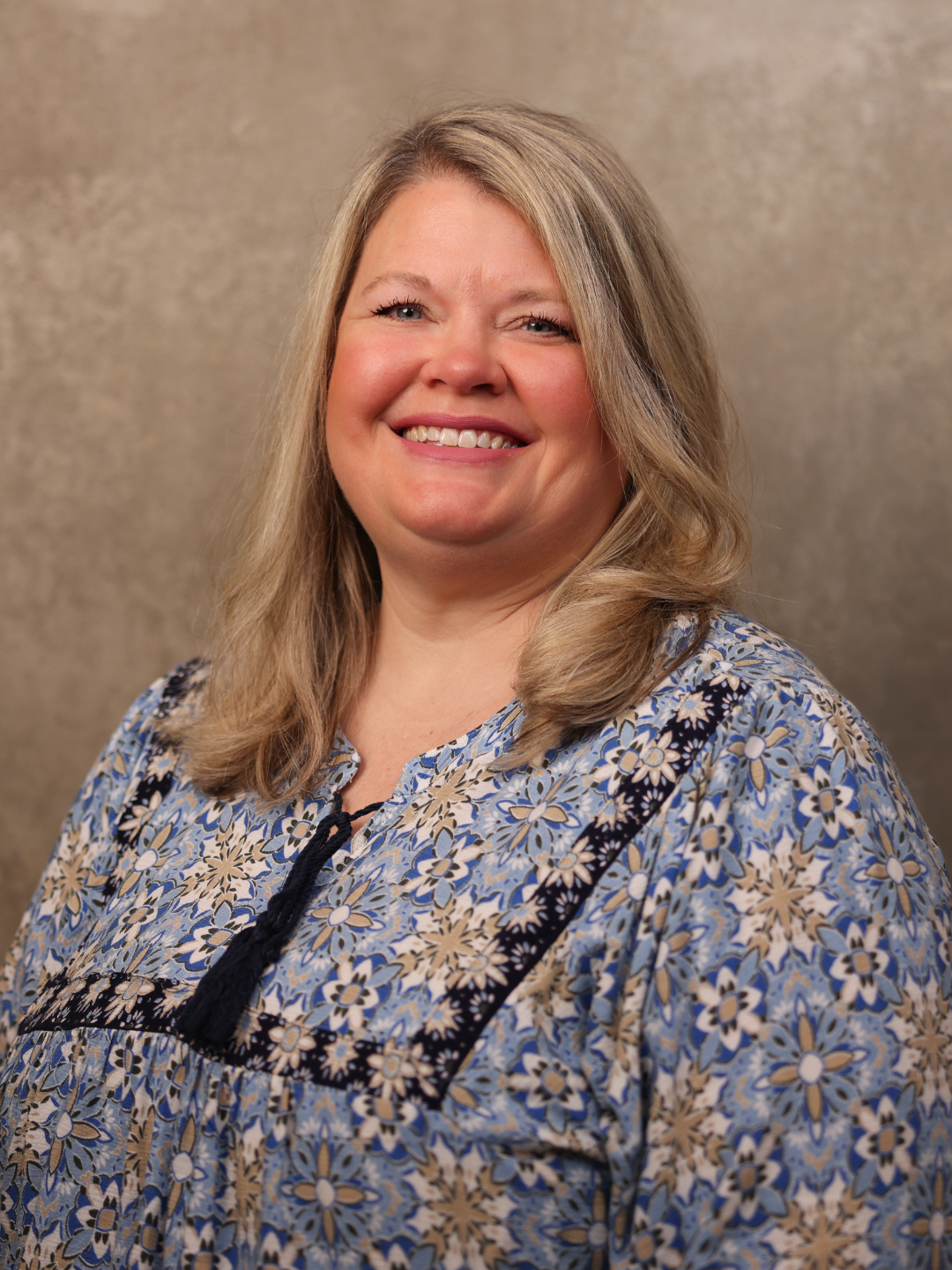 Portrait of a smiling woman with shoulder-length blonde hair, wearing a floral-patterned blouse, against a neutral background.