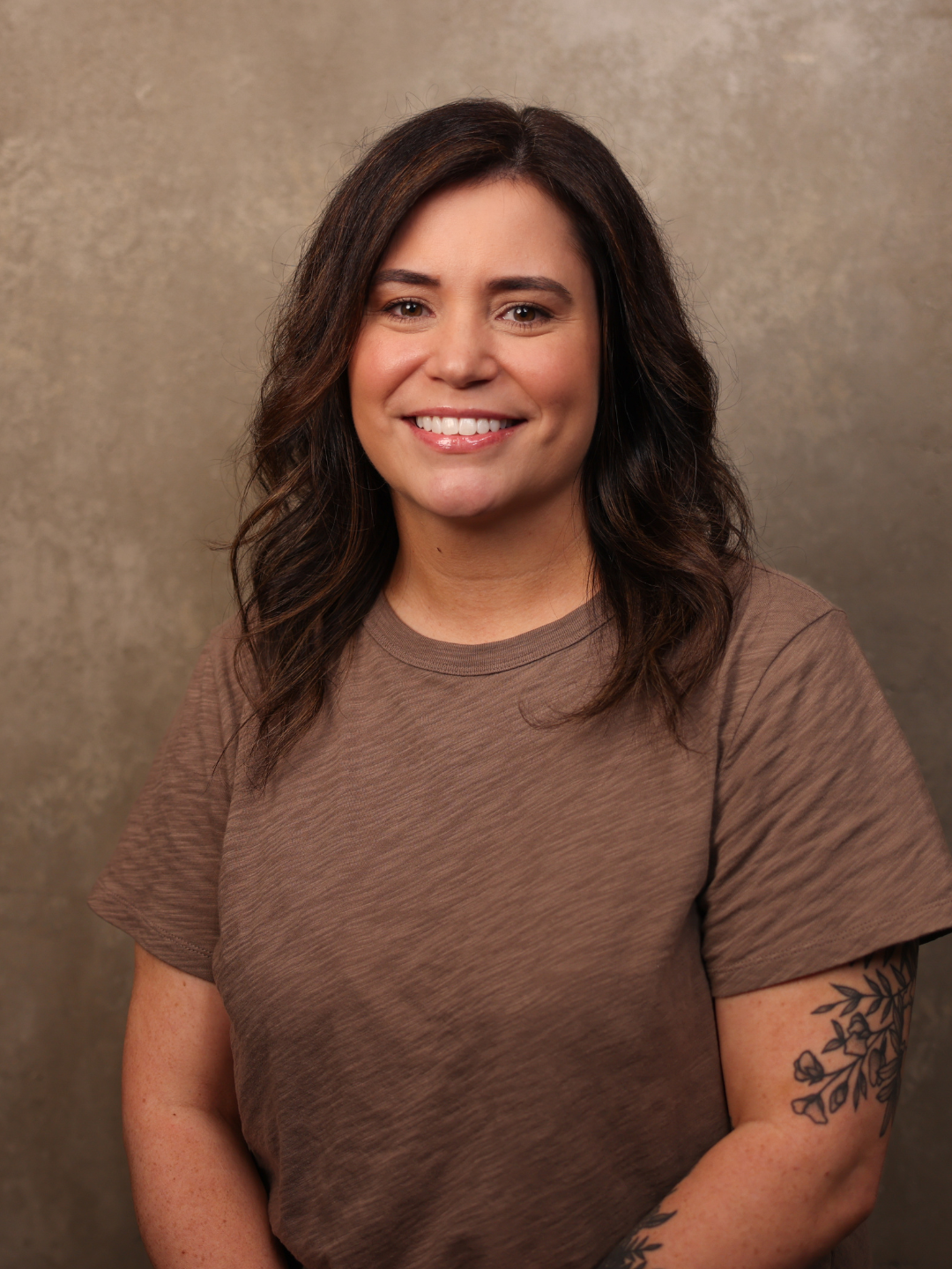 A woman with shoulder-length wavy brown hair, wearing a brown T-shirt, smiling in front of a textured beige background.