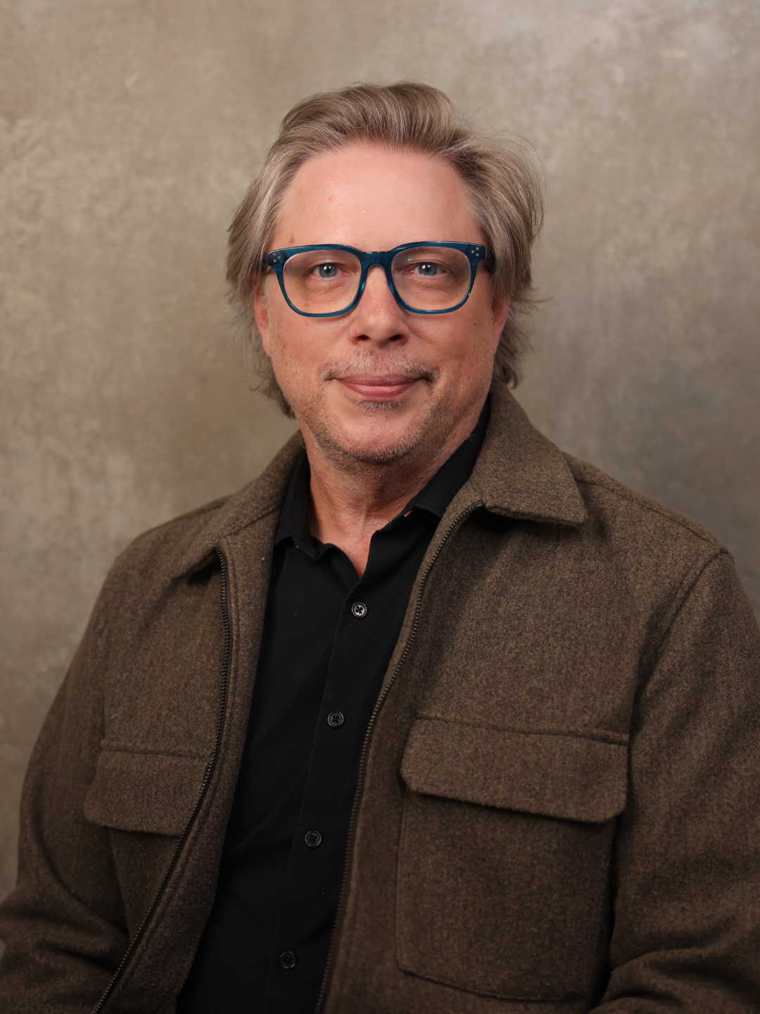A middle-aged man with glasses and gray hair, wearing a brown jacket and black shirt, smiling in front of a plain, textured background.