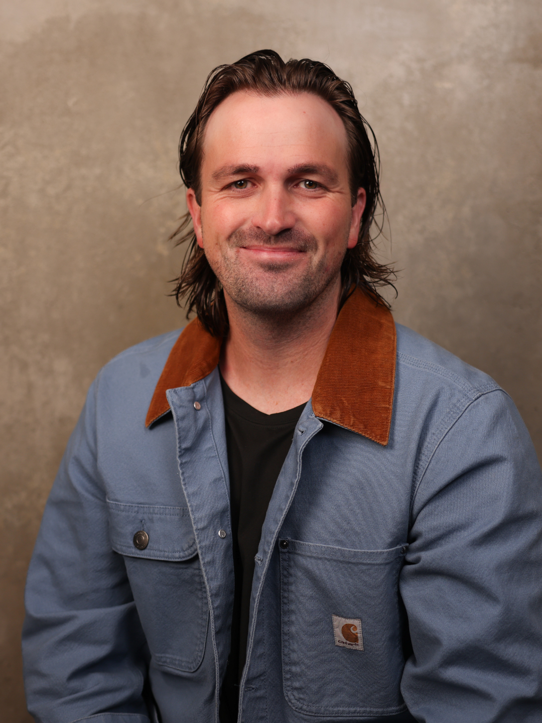 A man with shoulder-length brown hair, a slight beard, and a friendly smile, wearing a blue jacket with a brown collar and a black shirt underneath, seated against a plain, textured beige background.