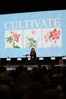 A woman standing on stage giving a presentation with a large screen displaying the word 'CULTIVATE' and images of flowers.