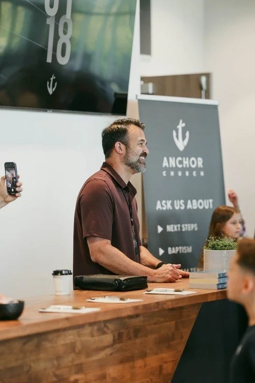 A man standing at a wooden countertop, smiling and speaking to an audience in a church or community center, with a banner that reads 'Anchor Church' in the background.