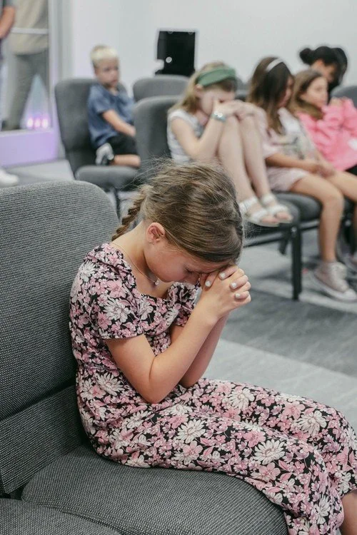 A girl in a floral dress praying with her hands clasped and head bowed, sitting on a gray chair in a room with other children, some sitting with heads bowed.