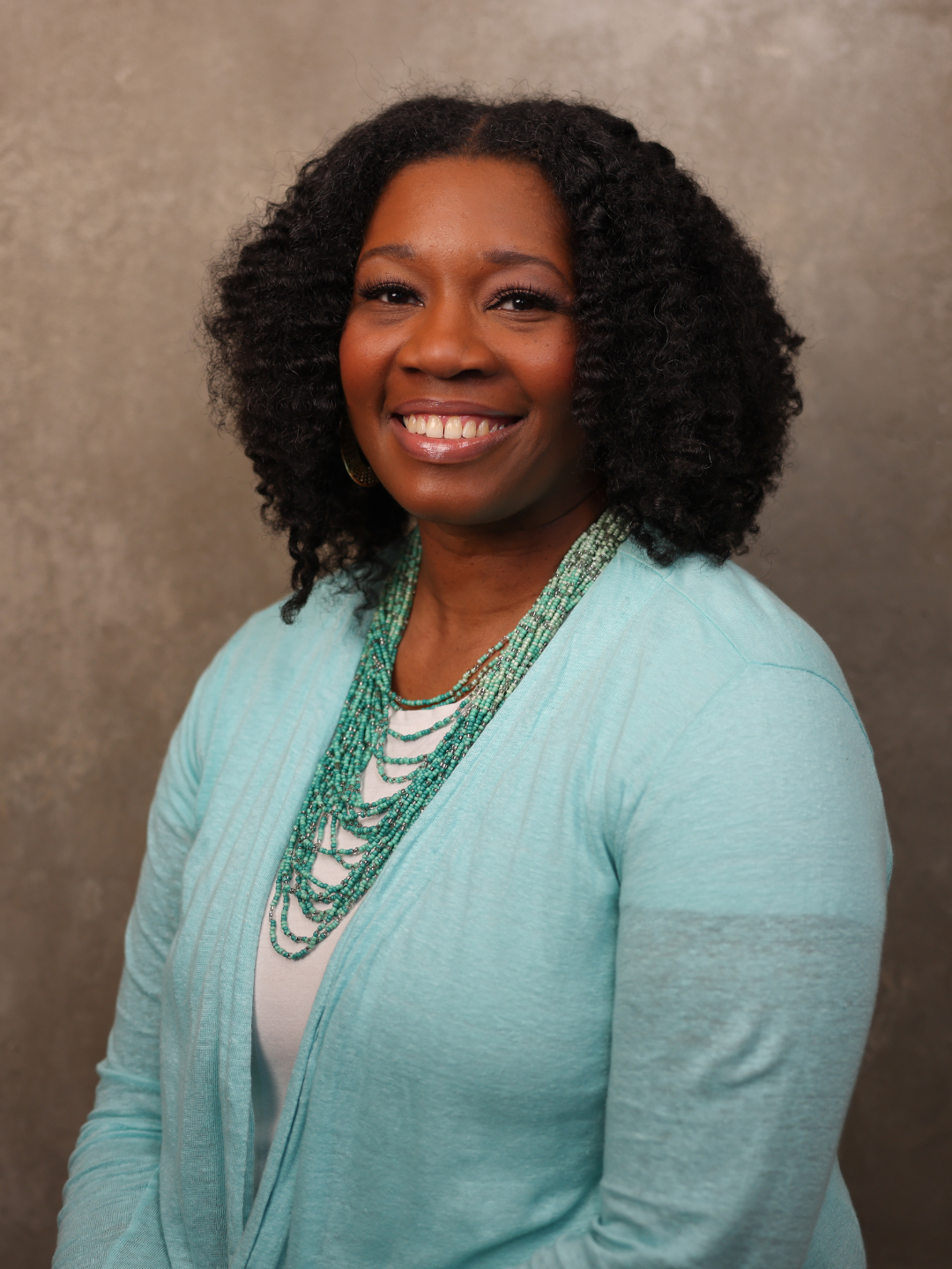 Portrait of a smiling African American woman with curly hair, wearing a light blue cardigan and layered turquoise necklaces.