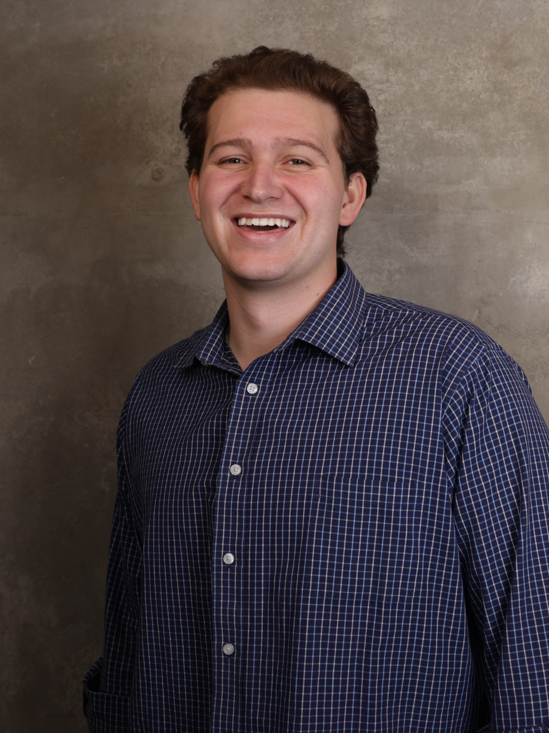 A young man with dark curly hair smiling, wearing a dark blue checkered shirt, standing in front of a textured gray wall.