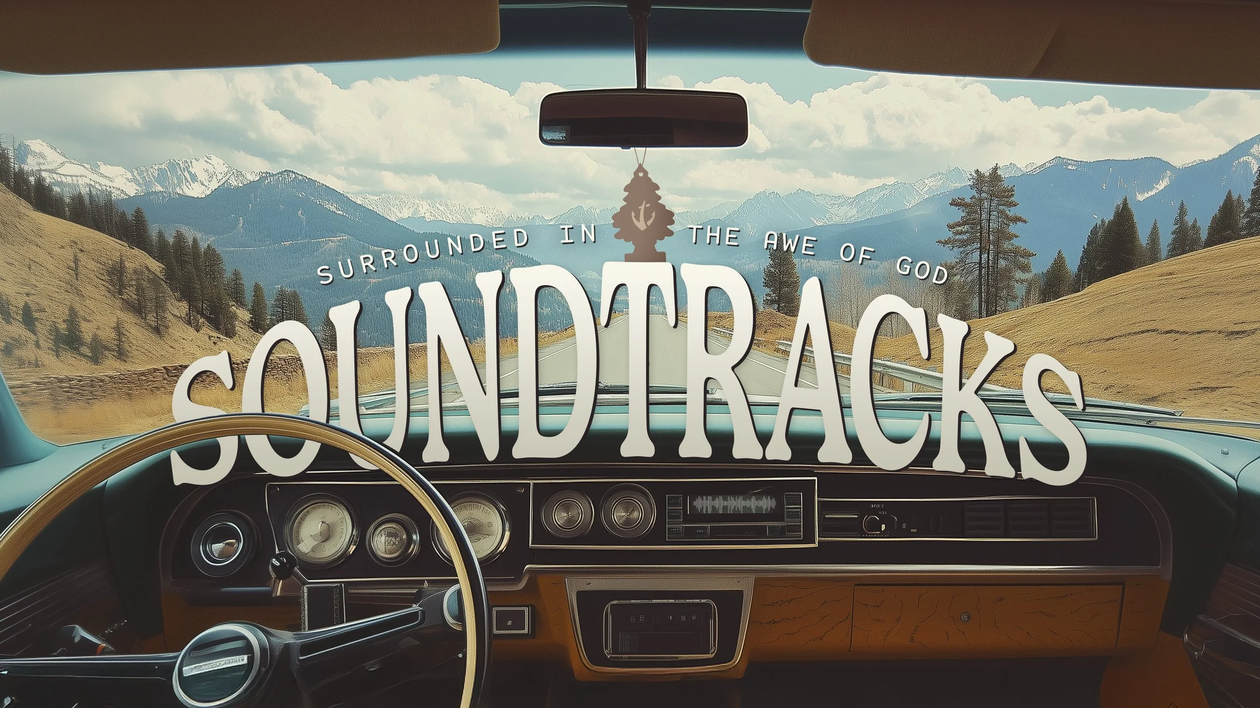The view from inside a vintage vehicle driving through a scenic mountainous landscape in South Dakotas, with mountains and trees visible through the windshield and a decorative hanging ornament from the rearview mirror.