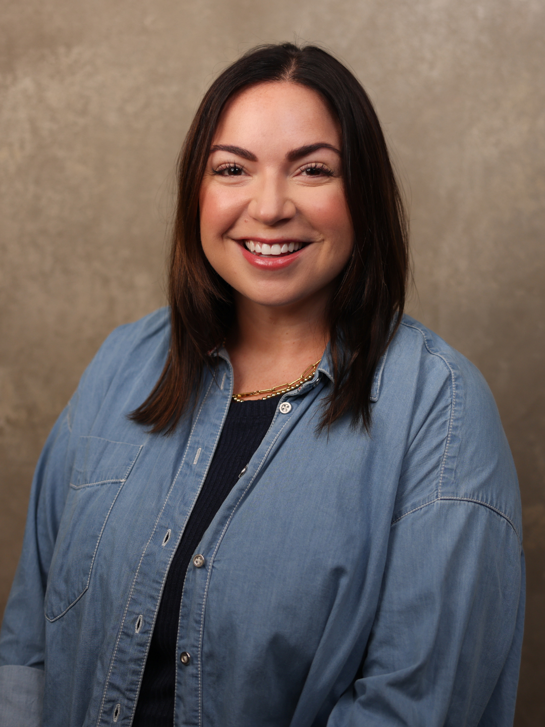 A woman with shoulder-length dark brown hair smiling, wearing a denim shirt and a gold necklace, against a neutral background.