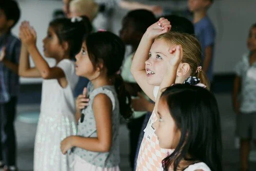 Children standing in a classroom, raising their hands and smiling.