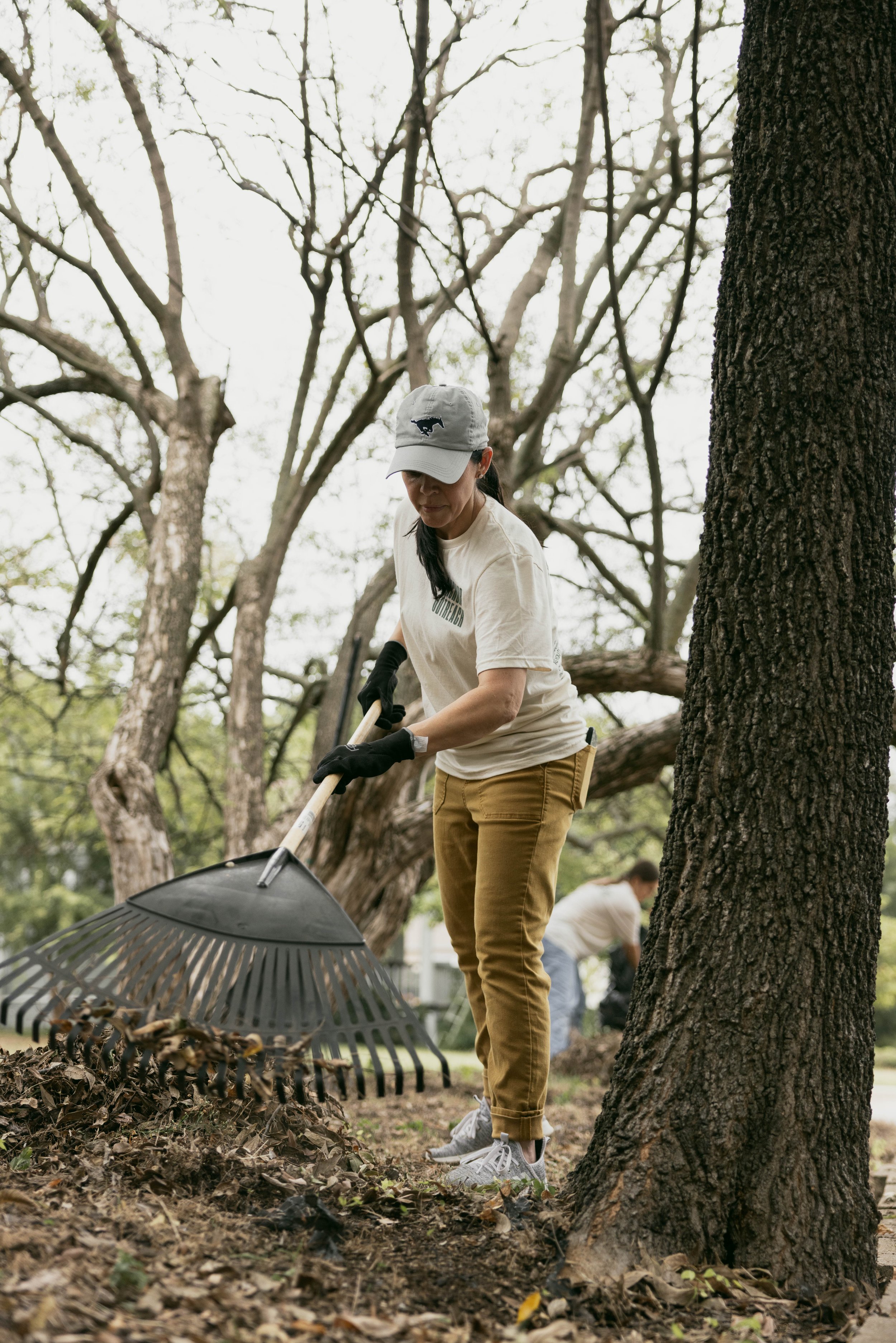 A woman raking leaves in a park or garden, wearing a light-colored T-shirt with a logo, yellow pants, and a light gray cap, with trees in the background.