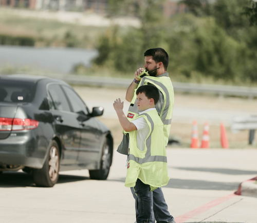 A man and a boy in safety vests are standing on the side of a road with cars passing by.