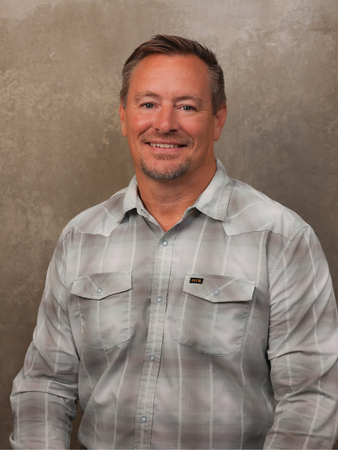A smiling middle-aged man with short brown hair, a goatee, and wearing a light gray plaid button-up shirt, standing against a plain, textured gray background.