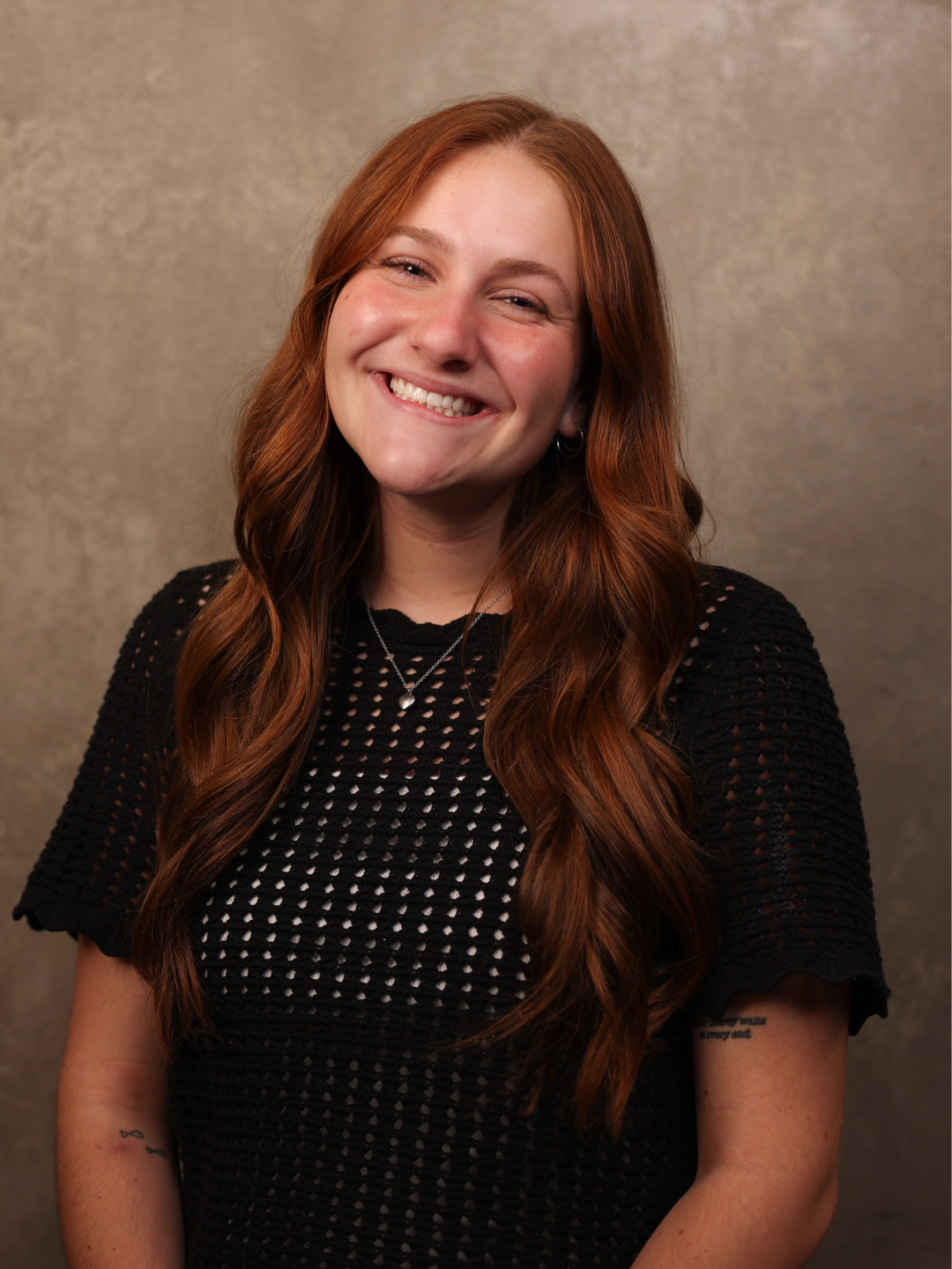 A young woman with long red hair, smiling and wearing a black textured top, standing against a beige background.