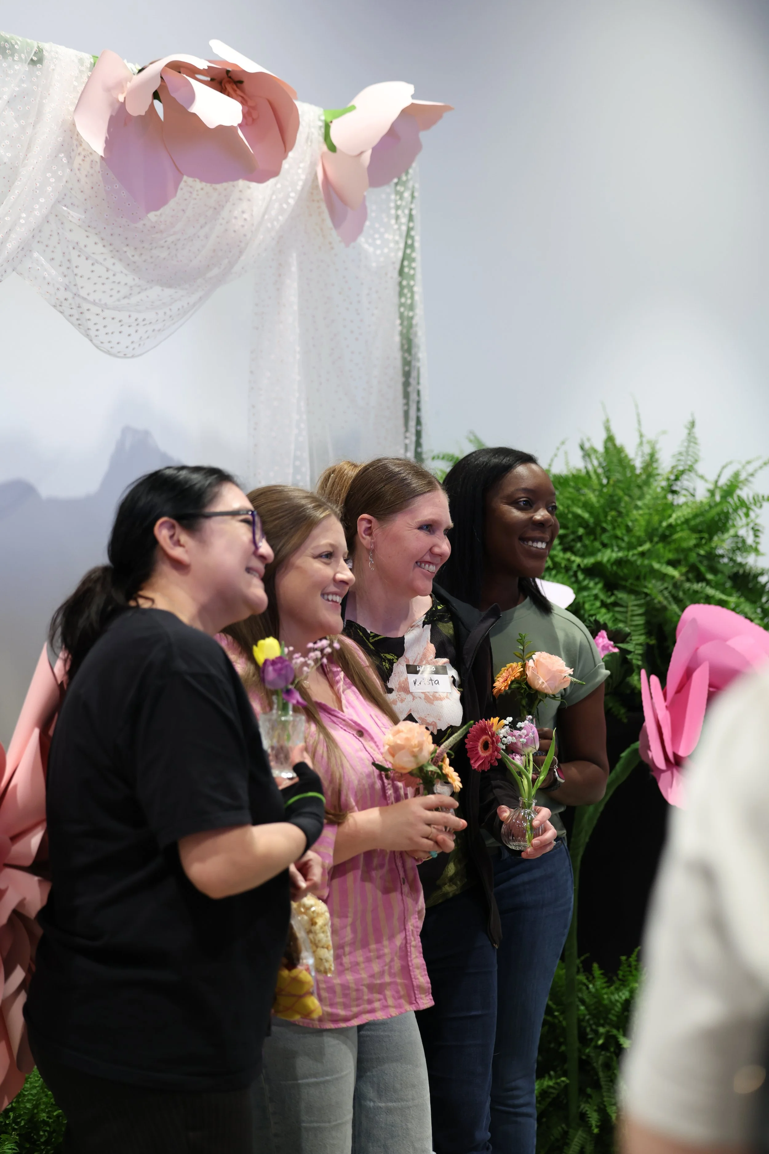 Group of diverse women smiling and holding flowers at an indoor event, with plants and decorative pink flowers in the background.