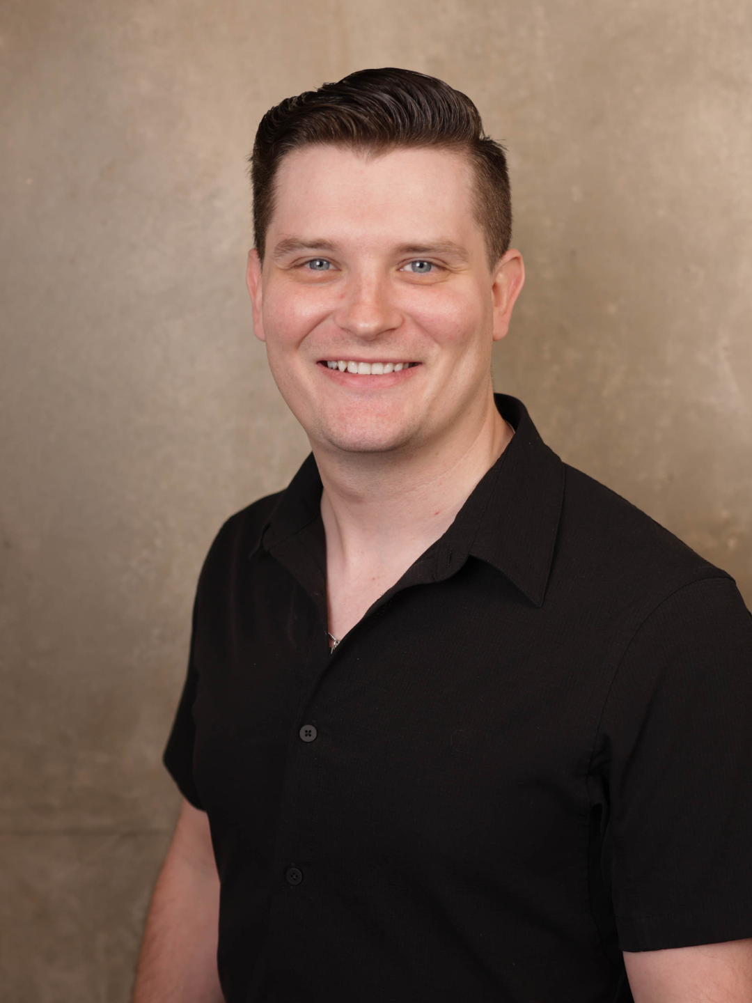 A portrait of a smiling man with short dark hair wearing a black collared shirt against a plain beige background.