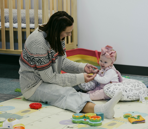 A woman playing with a baby girl on a colorful mat in a nursery room.
