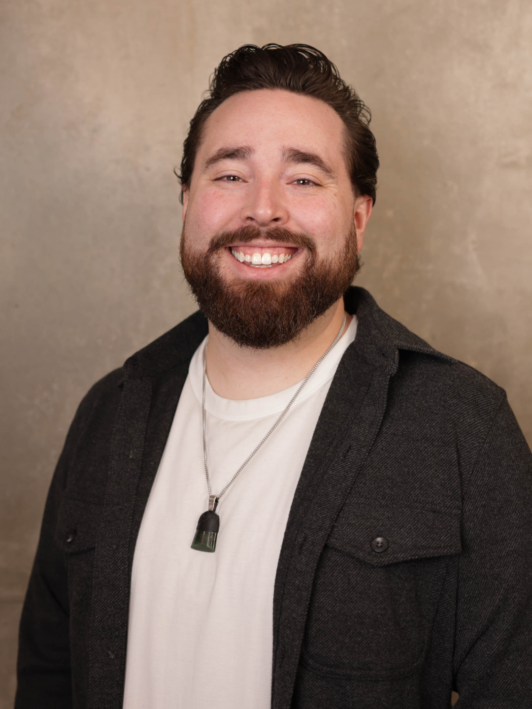 Portrait of a smiling man with dark hair, beard, wearing a white shirt, dark jacket, and a necklace with a black pendant, standing against a neutral background.
