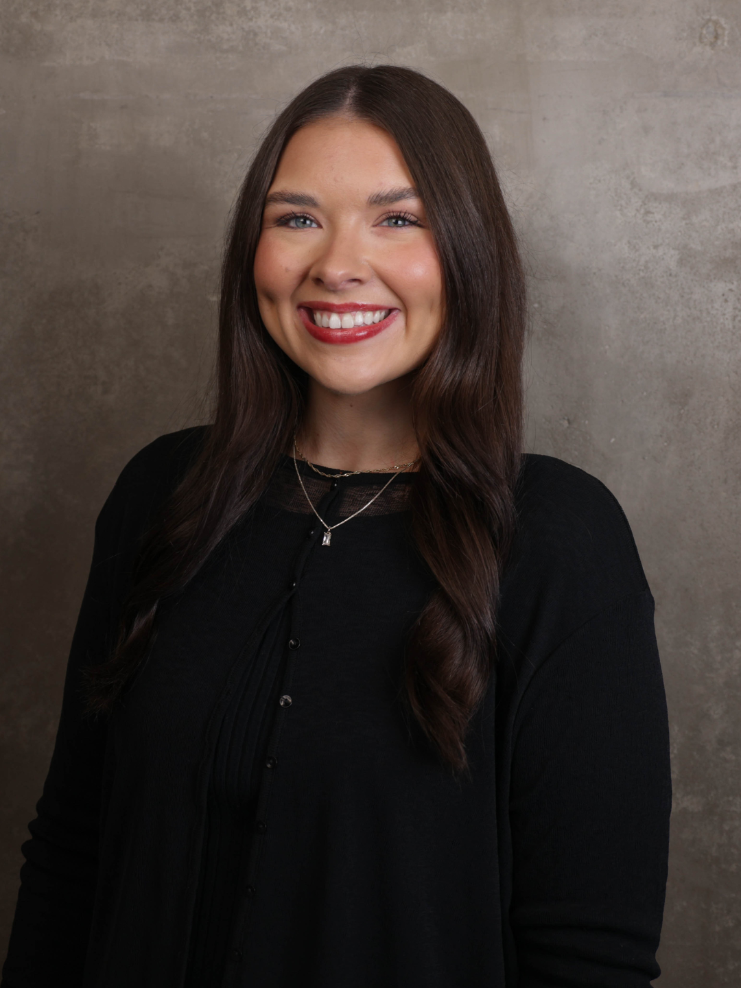 A woman with long brown hair, wearing a black top with buttons, smiling with braces, standing against a concrete background.