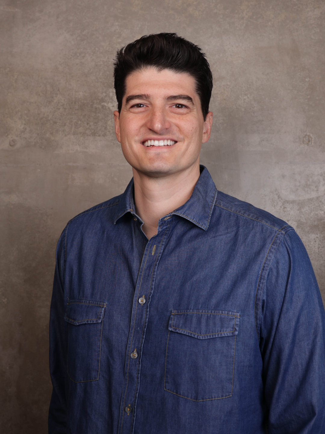 A man with dark hair, wearing a denim shirt, smiling in front of a plain, textured background.