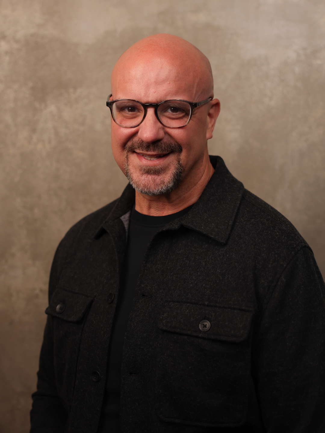 A middle-aged man with a bald head, glasses, and a goatee, smiling at the camera, wearing a dark button-up shirt over a black T-shirt, standing against a plain, beige background.