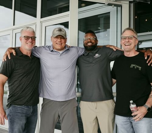 Four men standing outdoors, with arms around each other's shoulders, smiling for the camera in front of a glass building.