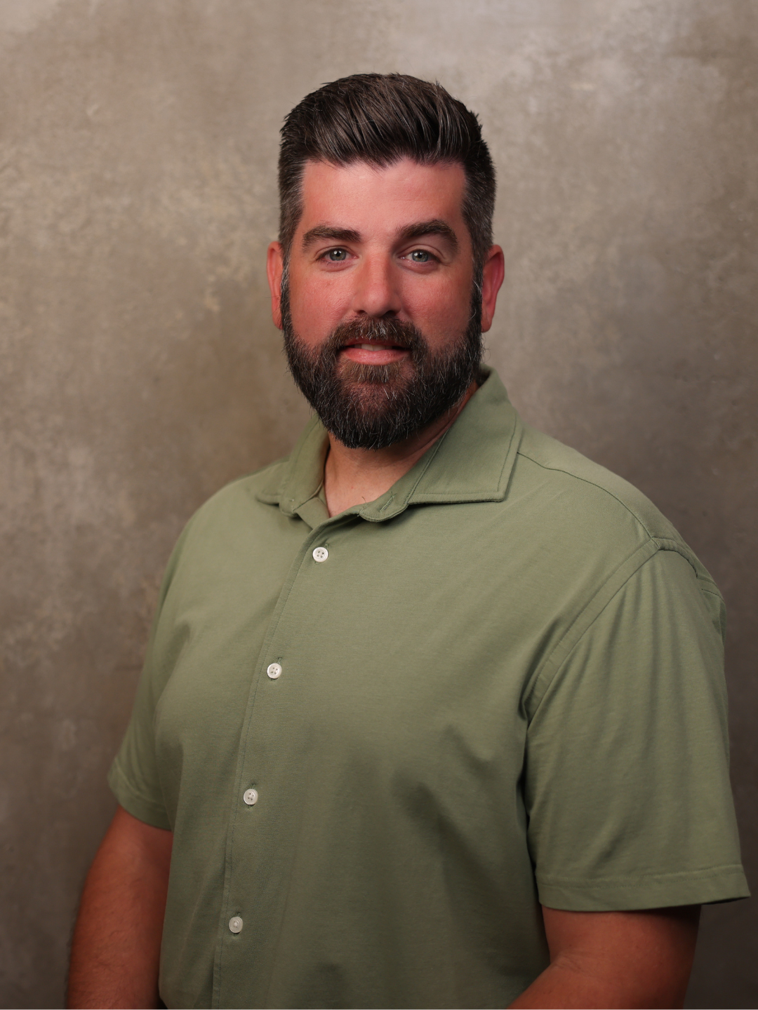 A headshot of a man with dark hair, a beard, and is wearing a green button-up shirt, standing against a neutral textured background.