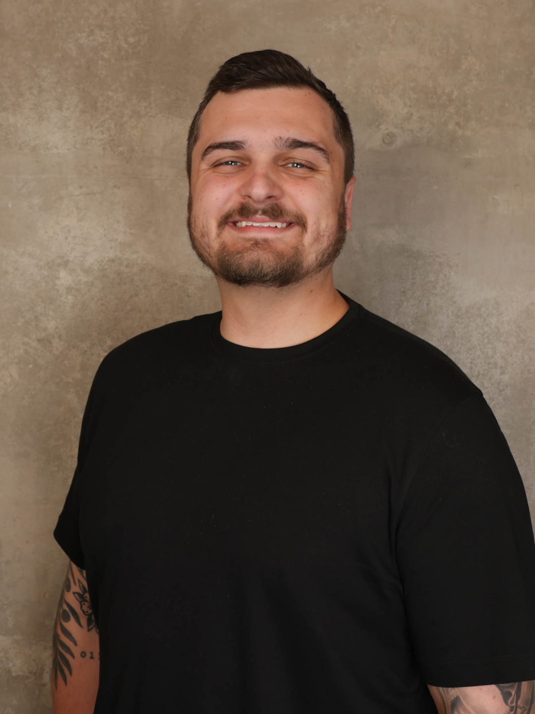 A smiling young man with short dark hair, a beard, and tattoos on his arm, wearing a black T-shirt, standing against a beige textured wall.