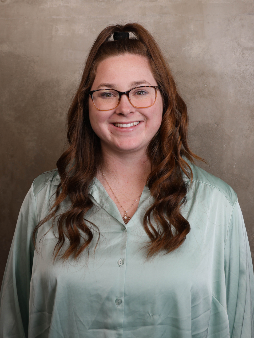 A woman with long wavy brown hair, glasses, and a light green button-up shirt, standing against a neutral textured background, smiling for the camera.