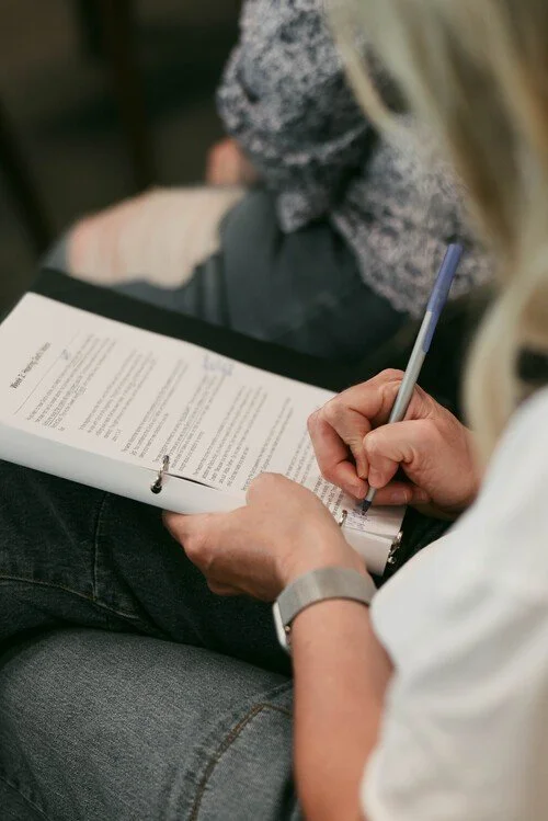 Person writing on a clipboard during a class or meeting, with other people sitting nearby.