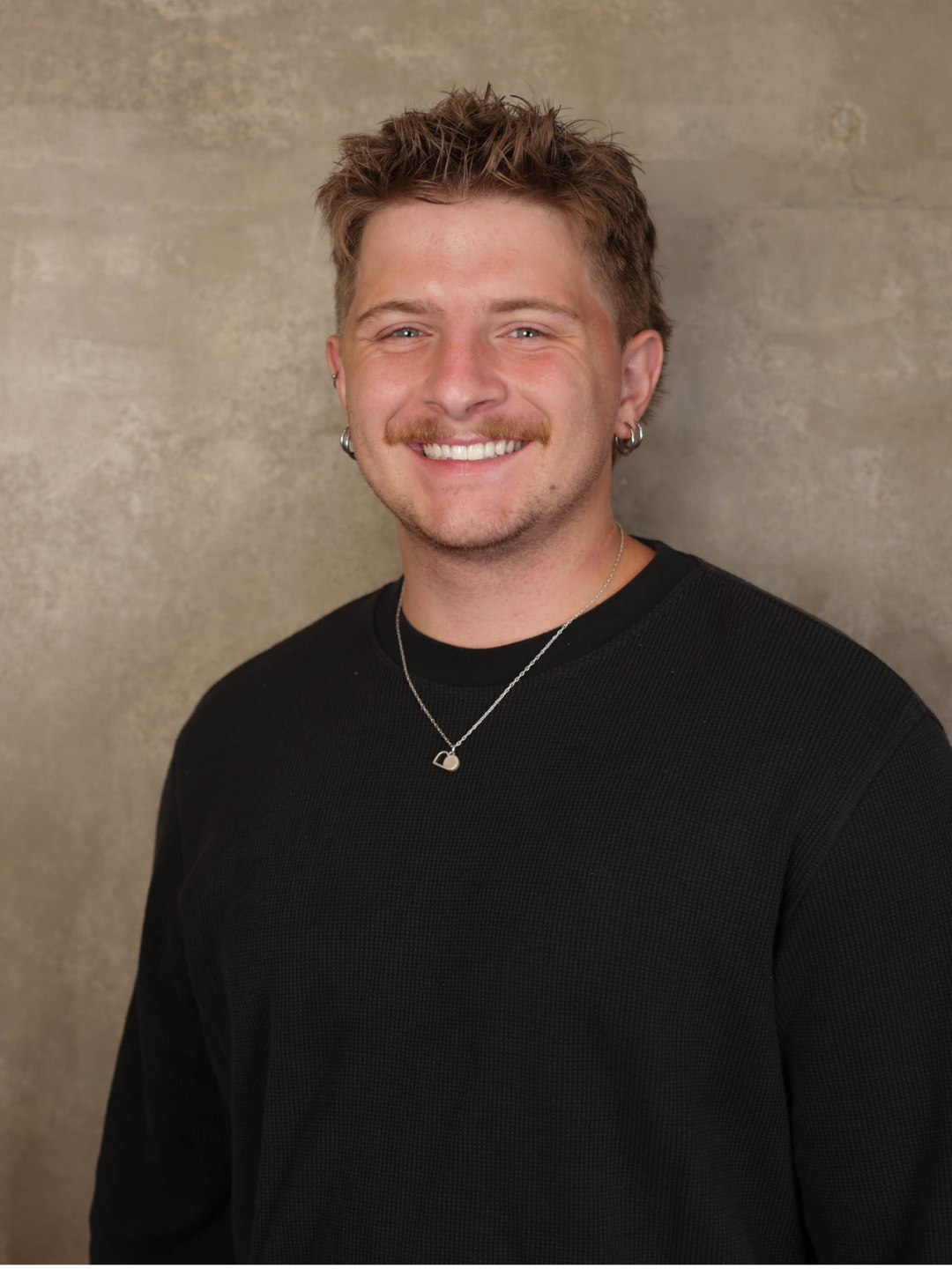 Smiling young man with short, spiky hair, wearing a black textured shirt, hoop earrings, a necklace with a heart pendant, and earrings, standing against a beige wall.