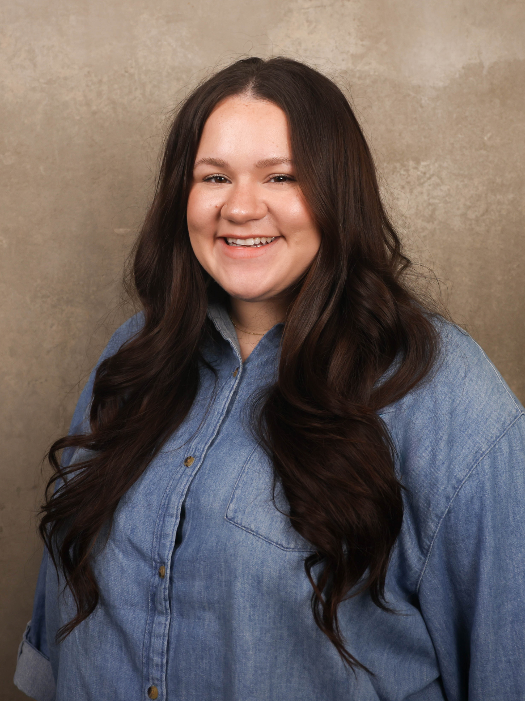 A young woman with long dark hair smiling, wearing a denim shirt, against a neutral background.
