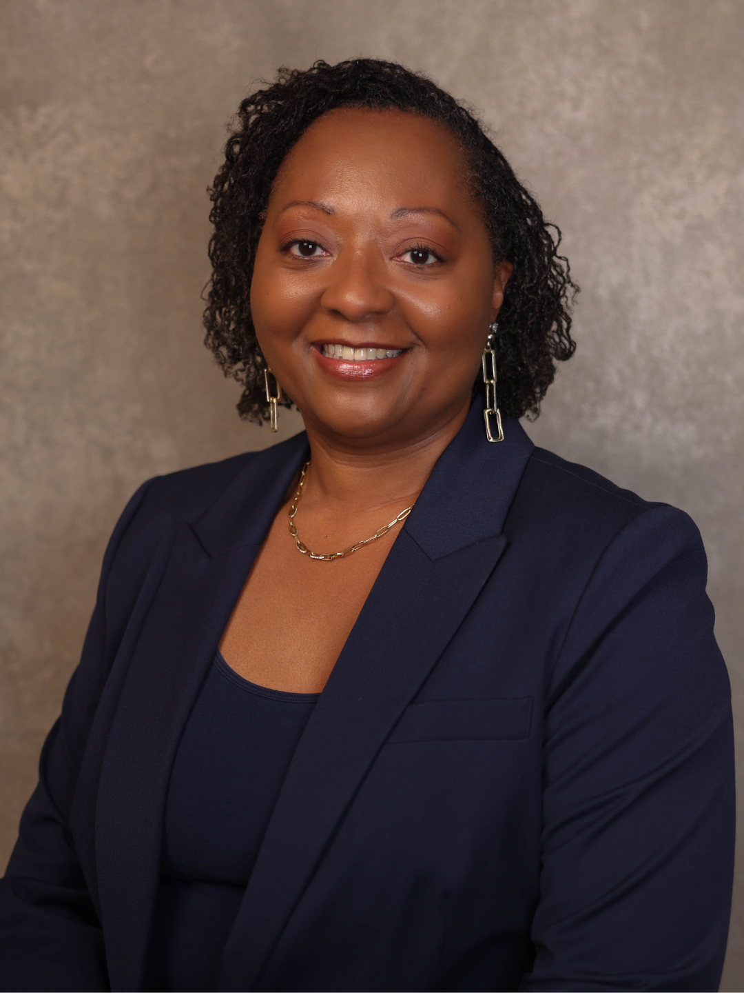 A professional woman with short curly hair wearing a navy blazer, matching top, and gold jewelry, smiling against a neutral background.
