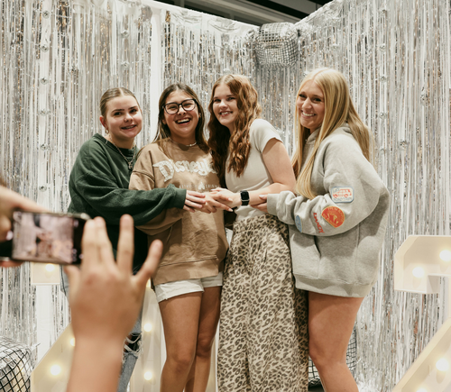 Four young women smiling and holding hands in front of a silver, sparkly backdrop, with someone taking their photo.