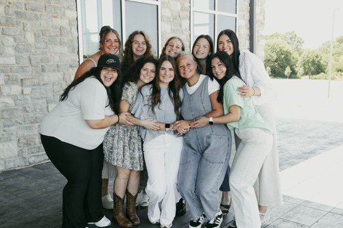 Group of nine smiling women standing together outside near a building, holding hands and posing for the photo.