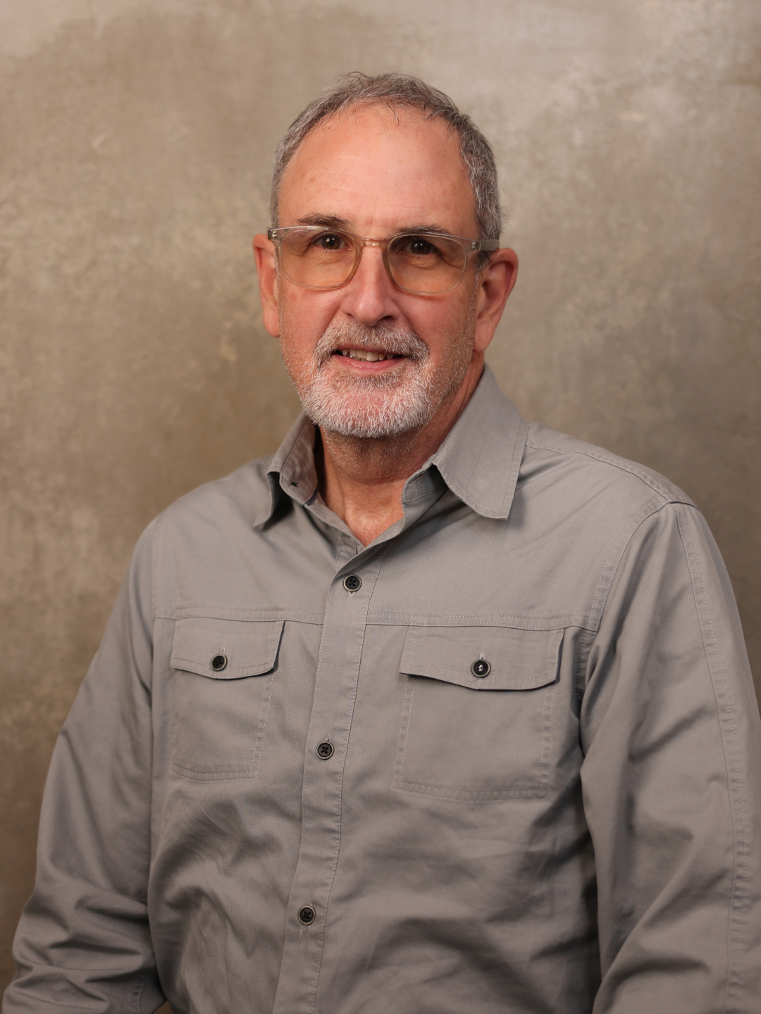 Portrait of an older man with glasses, gray hair, and a beard, wearing a gray button-up shirt, standing against a neutral background.