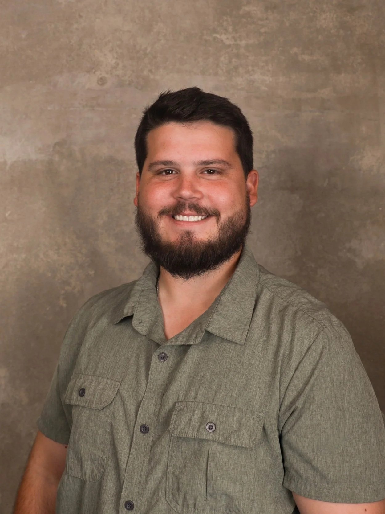 A smiling man with a beard, wearing a green button-up shirt, standing in front of a neutral textured background.