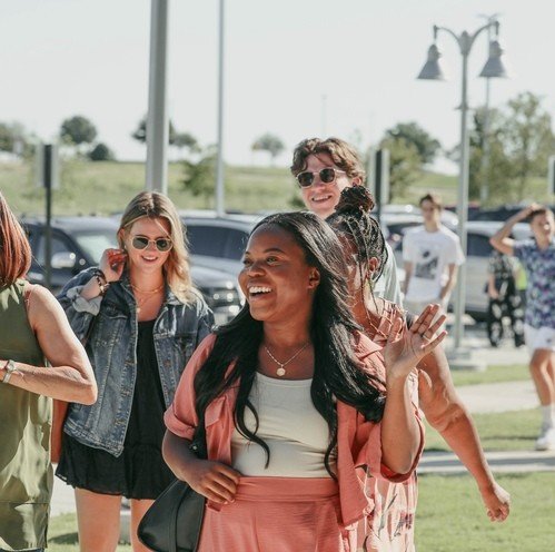 A group of smiling women walking outdoors on a sunny day, some wearing sunglasses, with cars and streetlights in the background.
